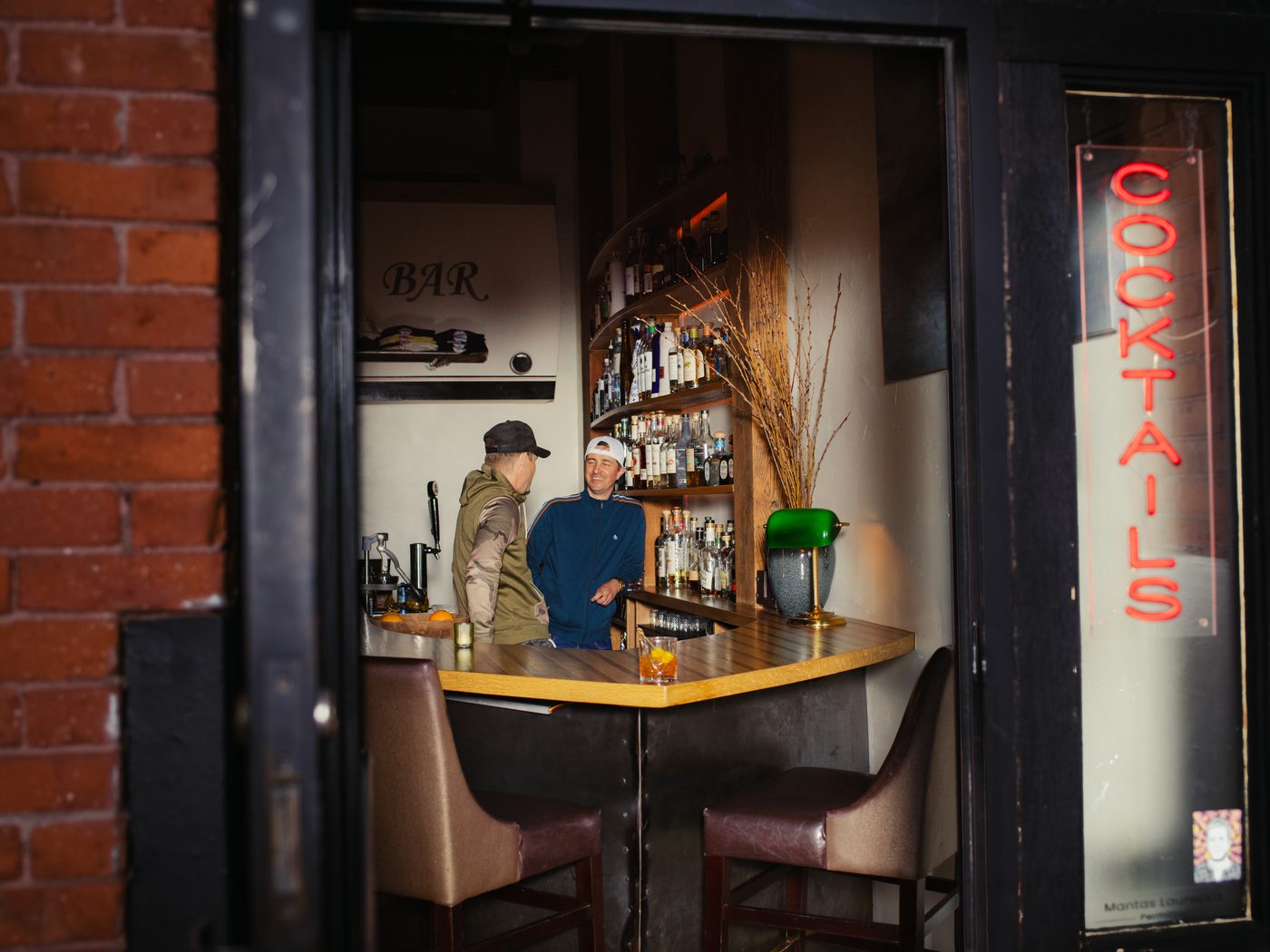 Looking through the doorway into Danger Bar: the back-bar shelves, a green banker's lamp, and guests talking, with a red 'Cocktails' neon at the entrance.